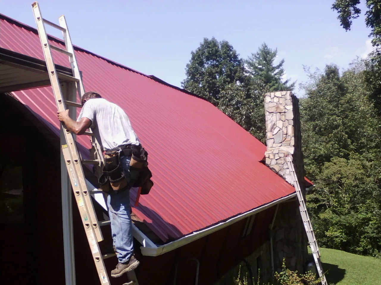Roofer on a ladder beside a red metal roof and stone chimney — Soffit & Fascia Repair by Roofing Brothers in Poland
